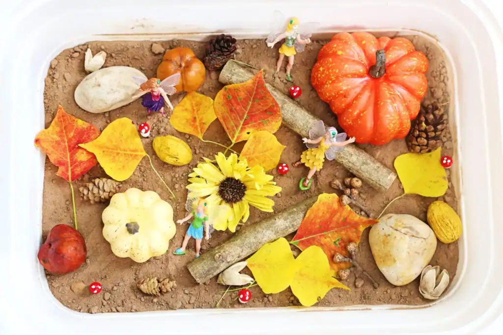 A fall sensory bin filled with brown sand, autumn leaves, pumpkins, pinecones, stones, toy fairies, and more—perfect for sensory play and kids activities inside a plastic container.