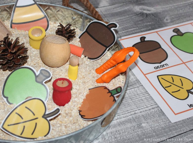 A fall sensory bin with paper cutouts of leaves and acorns, pinecones, a wooden cup, small figures, orange tongs, and a matching activity sheet offers an engaging sensory play experience for kids activities on a wooden surface.