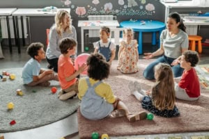 A group of young children sit in a circle on rugs in a classroom, while two adults guide them using techniques from an instructional strategies list; colorful toys and tables are visible in the background.