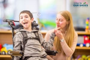 A young disabled child using a wheelchair smiles while a woman beside him holds his hand in a brightly lit indoor setting.
