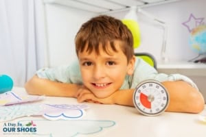 Smiling boy leans on a white desk with a visual timer and colorful paper cutouts, practicing time management in a bright, organized room.