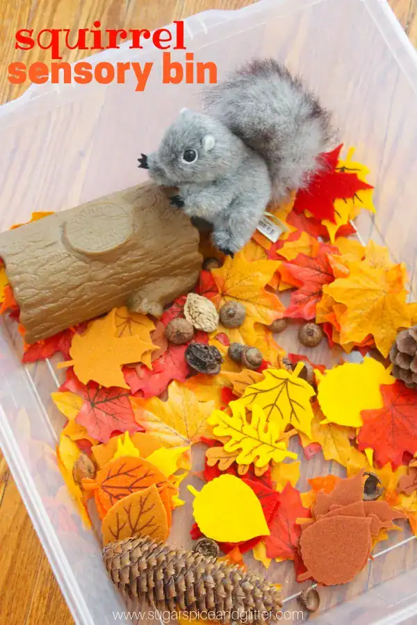 A clear plastic bin filled with artificial autumn leaves, acorns, pinecones, a toy log, and a plush squirrel creates an engaging fall sensory bin for kids to explore textures and ignite imaginative play.