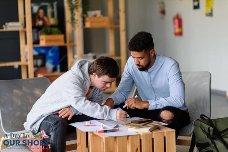 Two young men sit at a table working together on paperwork and drawings related to special needs education, with notebooks, papers, and pens spread out in front of them in a casual indoor setting.