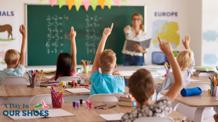 Children seated at desks in a classroom raise their hands while a teacher stands at the chalkboard with math problems written on it. Classroom decorations and maps are visible, supporting iep goals and supplemental educational services.