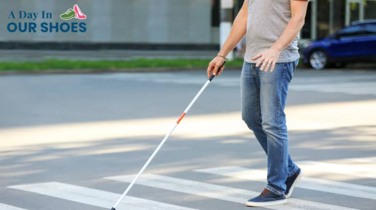 A person wearing jeans and a gray shirt walks across a crosswalk using a white cane, highlighting visual impairment and the need for transition assessments for students with blindness or severe disabilities. A blue car and building are in the background.