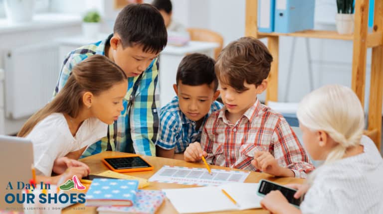 Five children sit around a table, examining a paper chart and using tablets and calculators in a classroom setting, exploring a sample giep. The "a day in our shoes" logo appears in the bottom left corner.