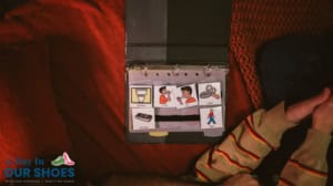 A communication binder used in iep assessments for non-verbal students, with illustrated cards for "bathroom," "drink," "sock," "keyboard," and "walk," is open on a couch beside a person in a striped shirt.