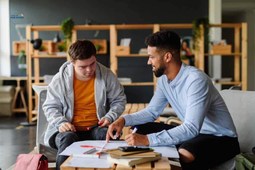 Two young men sit at a table with papers and notebooks, engaged in a discussion or tutoring session focused on re evaluation for IEP eligibility determination for students aged 18-21 in a modern, casual workspace.