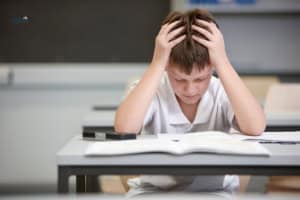 A boy in a white shirt sits at a desk in a classroom, holding his head in his hands while looking down at an open book, showing signs of adverse educational impact.