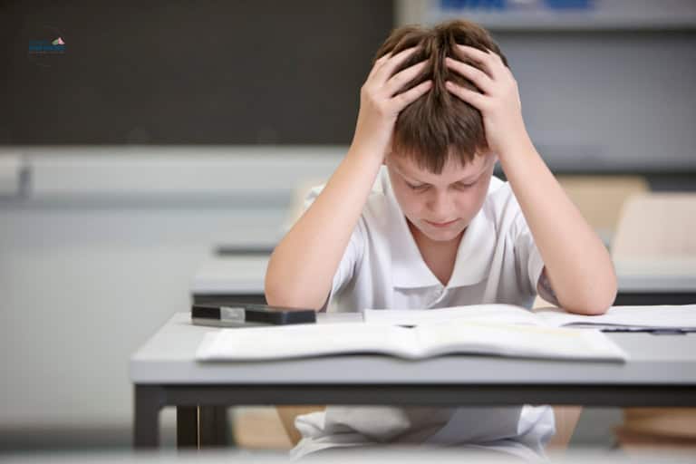 A boy in a white shirt sits at a desk in a classroom, holding his head in his hands while looking down at an open book, showing signs of adverse educational impact.