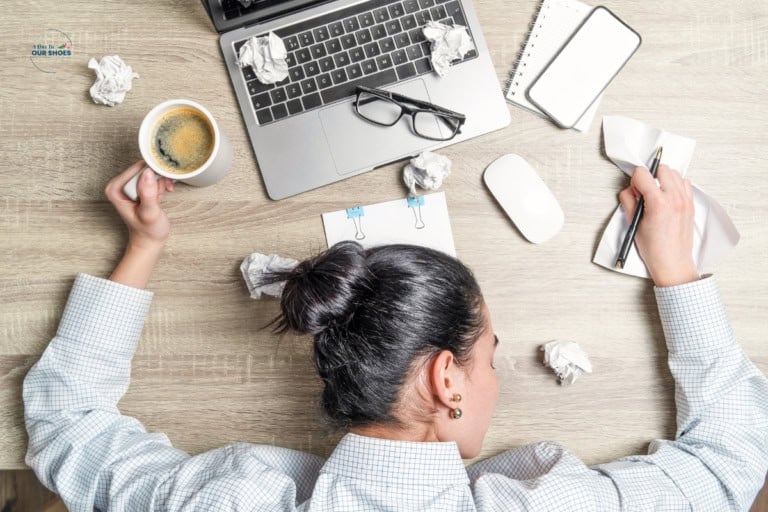 A person with their head down on a desk near a laptop, coffee, glasses, mouse, papers, and crumpled paper balls, appearing exhausted—highlighting the need for executive function accommodations in the workplace.