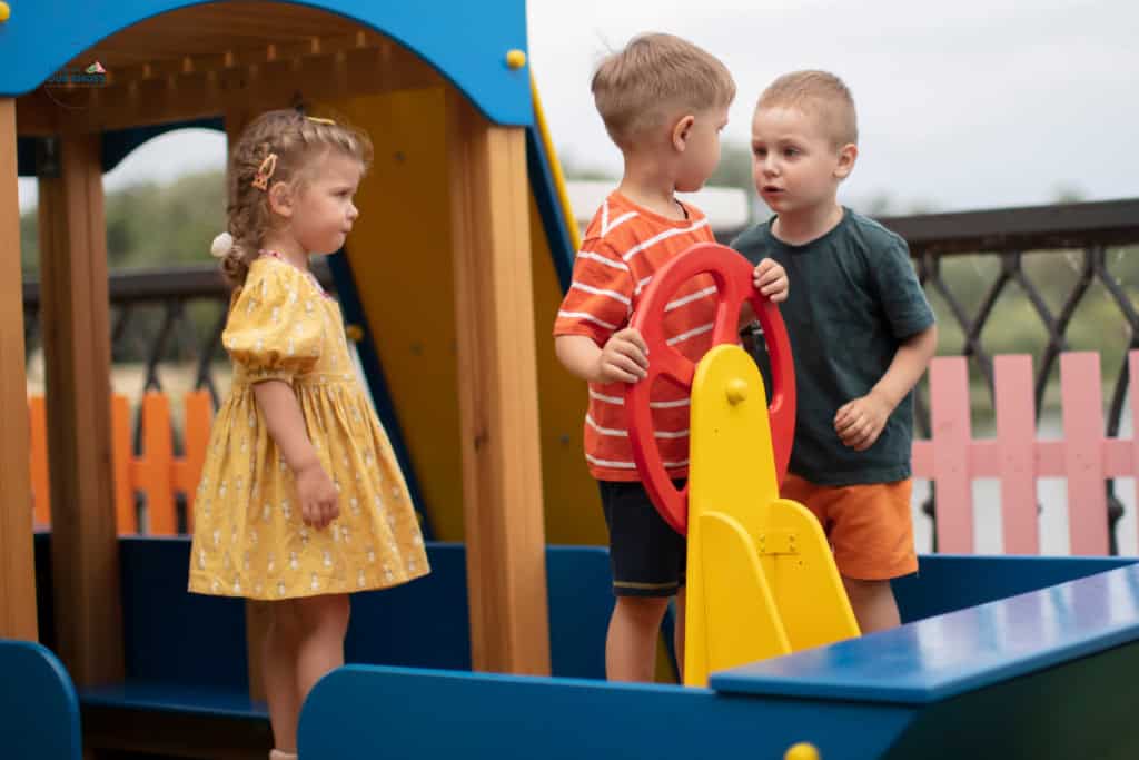 Three young children play together on a colorful playground structure, with two boys at a steering wheel and a girl standing nearby watching them, naturally teaching pragmatics through their playful interactions.