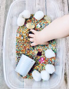 A child's hand in a st. Patrick's day sensory bin container with sprinkles in it.